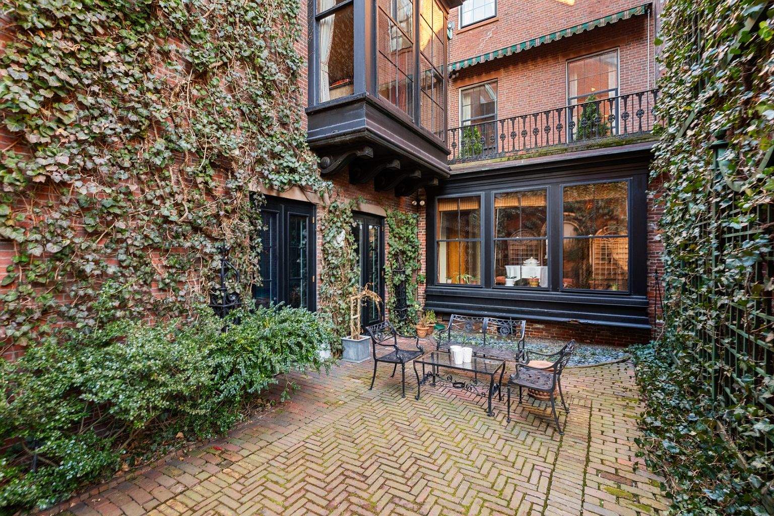 This image showcases a charming brick patio area, enclosed by brick walls partially covered in lush ivy. A wrought-iron table and chairs set provides a cozy seating arrangement, suggesting an inviting outdoor space for relaxation or entertaining. The architectural details, including the dark-framed windows and balcony above, add to the property's character and appeal.