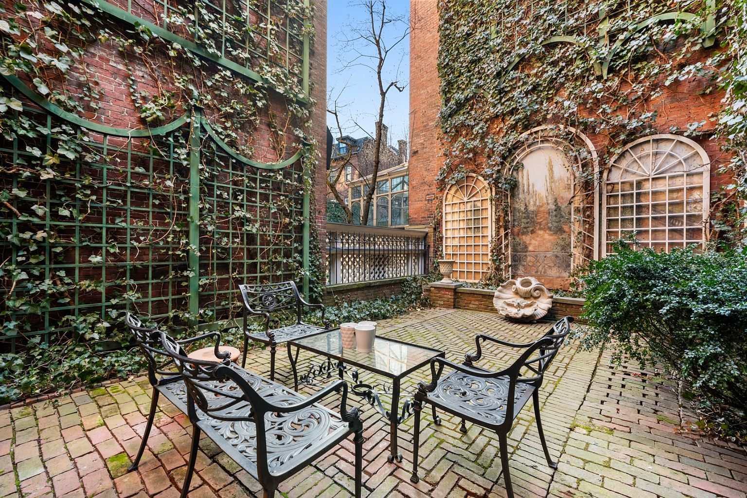 This image showcases a charming outdoor patio area, featuring a brick-paved surface and surrounded by brick walls covered in climbing ivy. A wrought-iron table and chairs set provides a cozy seating arrangement, complemented by decorative architectural elements like arched windows and a sculpted wall feature. The space exudes a sense of secluded elegance and old-world charm.