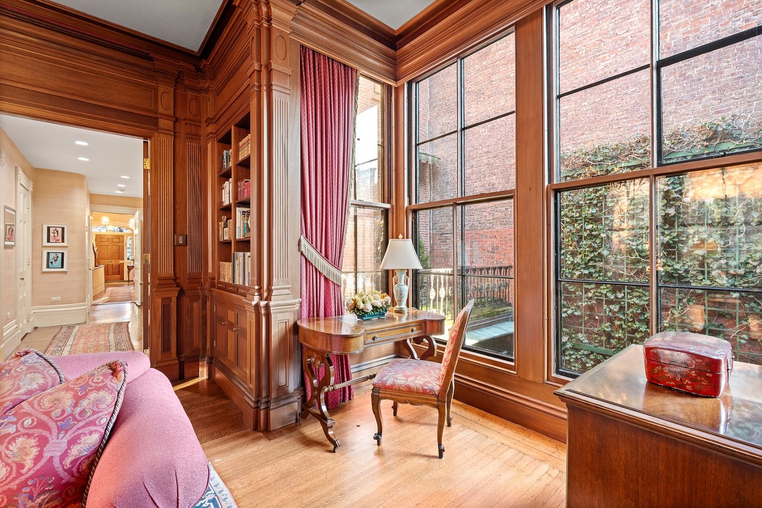 This is an interior shot of a home office or study, featuring rich wood paneling and built-in bookshelves. A writing desk sits in front of a large window offering natural light and a view of a brick wall covered in ivy. The room exudes a sense of classic elegance and sophistication.