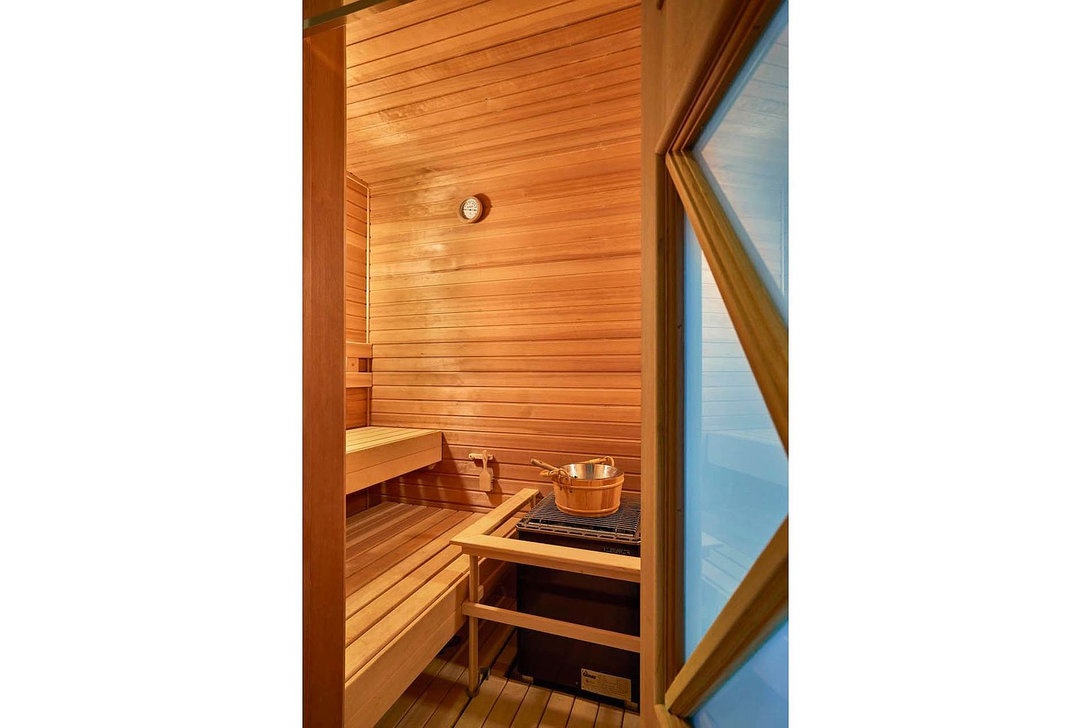 This image showcases a well-maintained sauna, featuring wooden benches and walls that create a warm and inviting atmosphere. A traditional sauna bucket and ladle sit atop the heater, adding to the authentic experience. The room is illuminated with soft lighting, enhancing the natural wood grain and promoting relaxation.