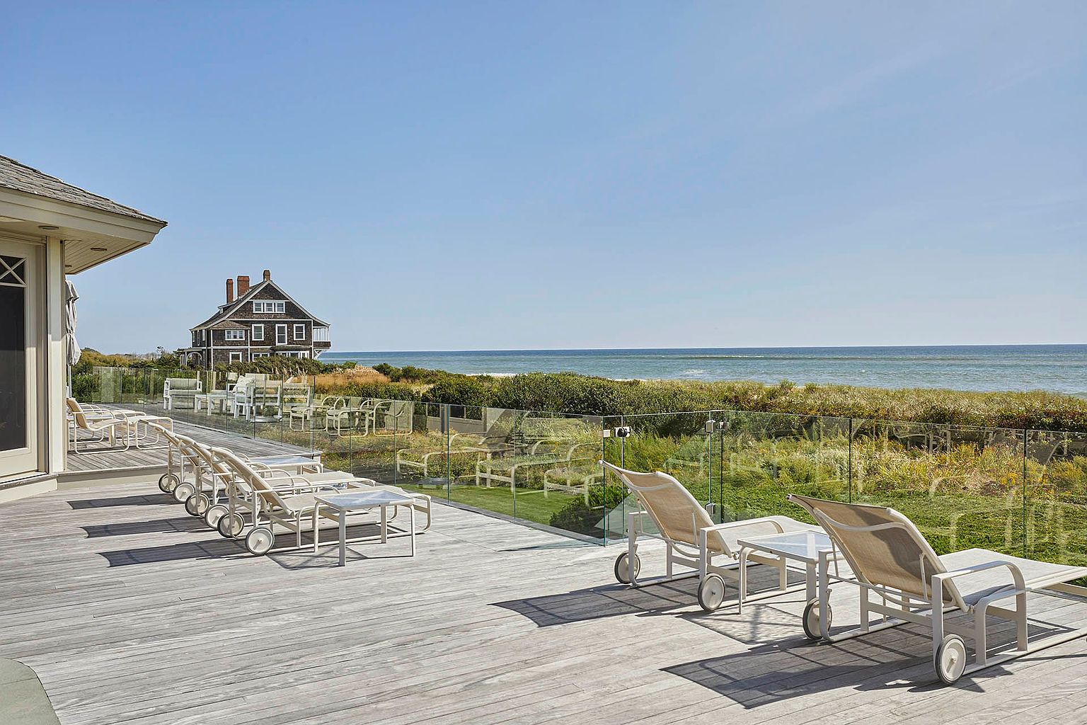 This image showcases a spacious wooden deck with multiple lounge chairs and small tables, offering a relaxing outdoor living space. A glass railing provides an unobstructed view of the ocean and surrounding landscape, while a charming house is visible in the background. The scene evokes a sense of luxury and tranquility, perfect for enjoying coastal living.
