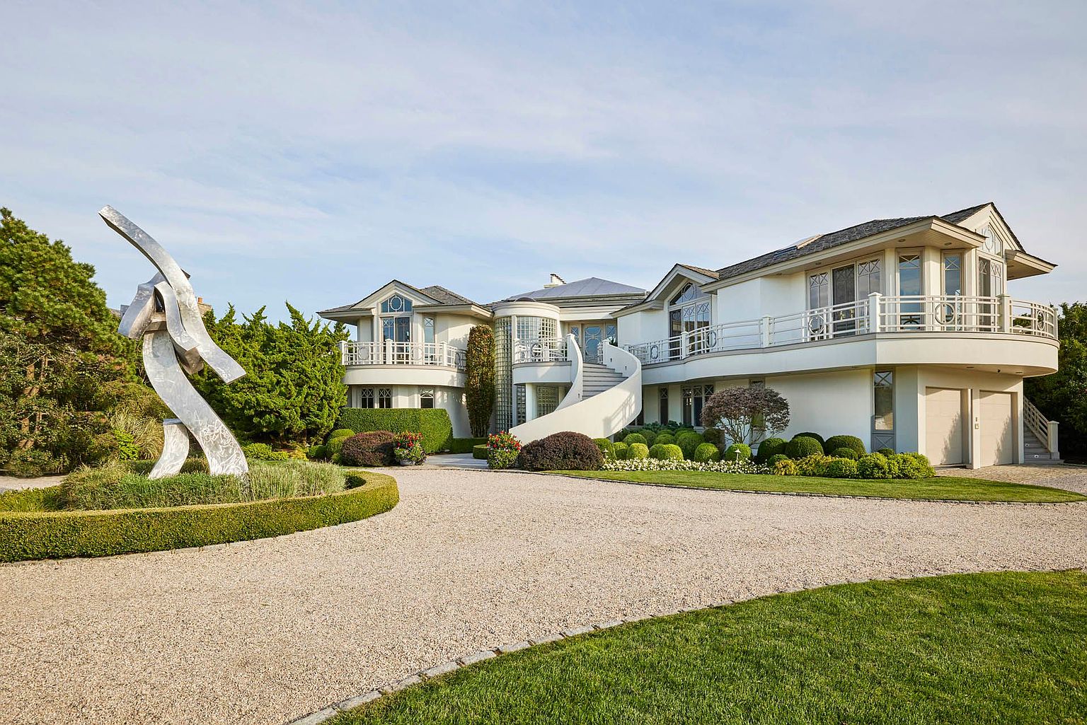 This is a front exterior view of a luxurious, modern home featuring a circular driveway, manicured landscaping, and a striking metal sculpture. The house is painted white and has multiple balconies with decorative railings, a curved staircase, and a unique architectural design. The overall impression is one of elegance and sophistication.