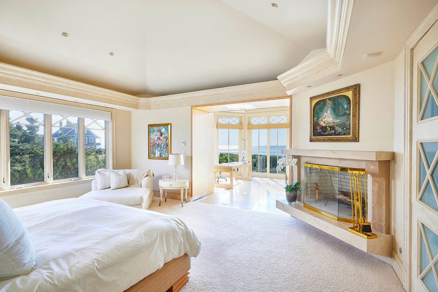 This is a luxurious primary bedroom featuring a large bed with white linens, a seating area by a window with views of greenery, and a fireplace with a decorative mantel. The room has an open layout with a separate office space visible in the background, and the overall design is elegant and bright with neutral tones and high-end finishes. The room is spacious and well-lit, creating a serene and inviting atmosphere.