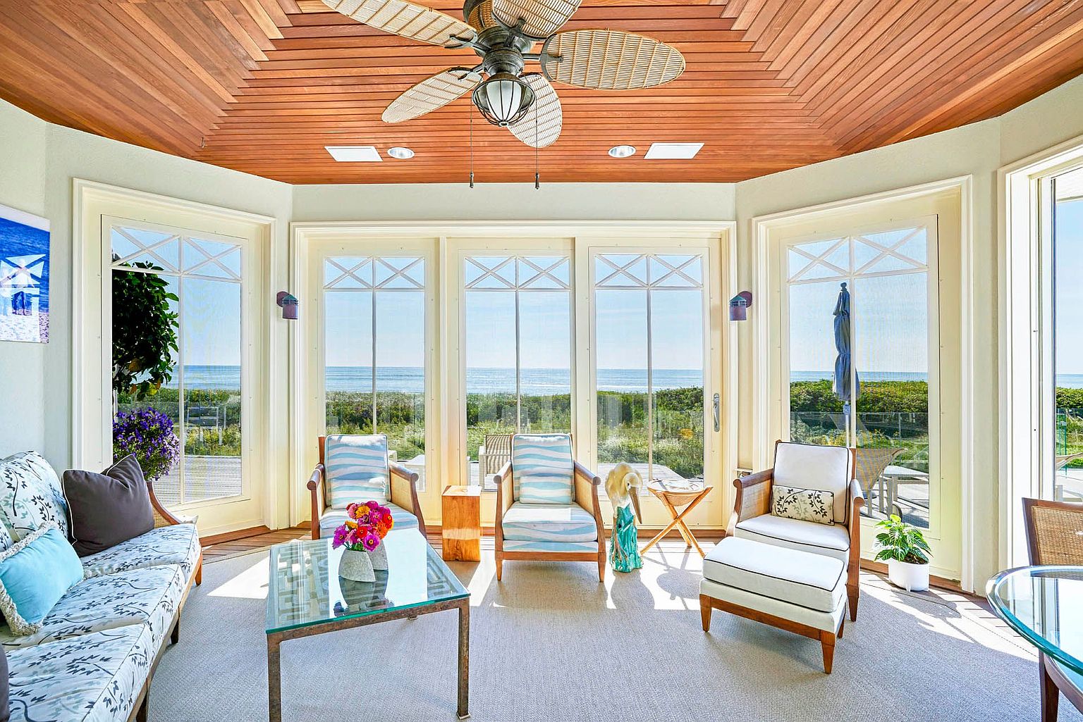 This sunroom features a wood-paneled ceiling with a ceiling fan and recessed lighting. Large windows offer a panoramic view of the ocean and surrounding greenery. The room is furnished with a sofa, chairs, a glass coffee table, and various decorative items, creating a bright and inviting space.