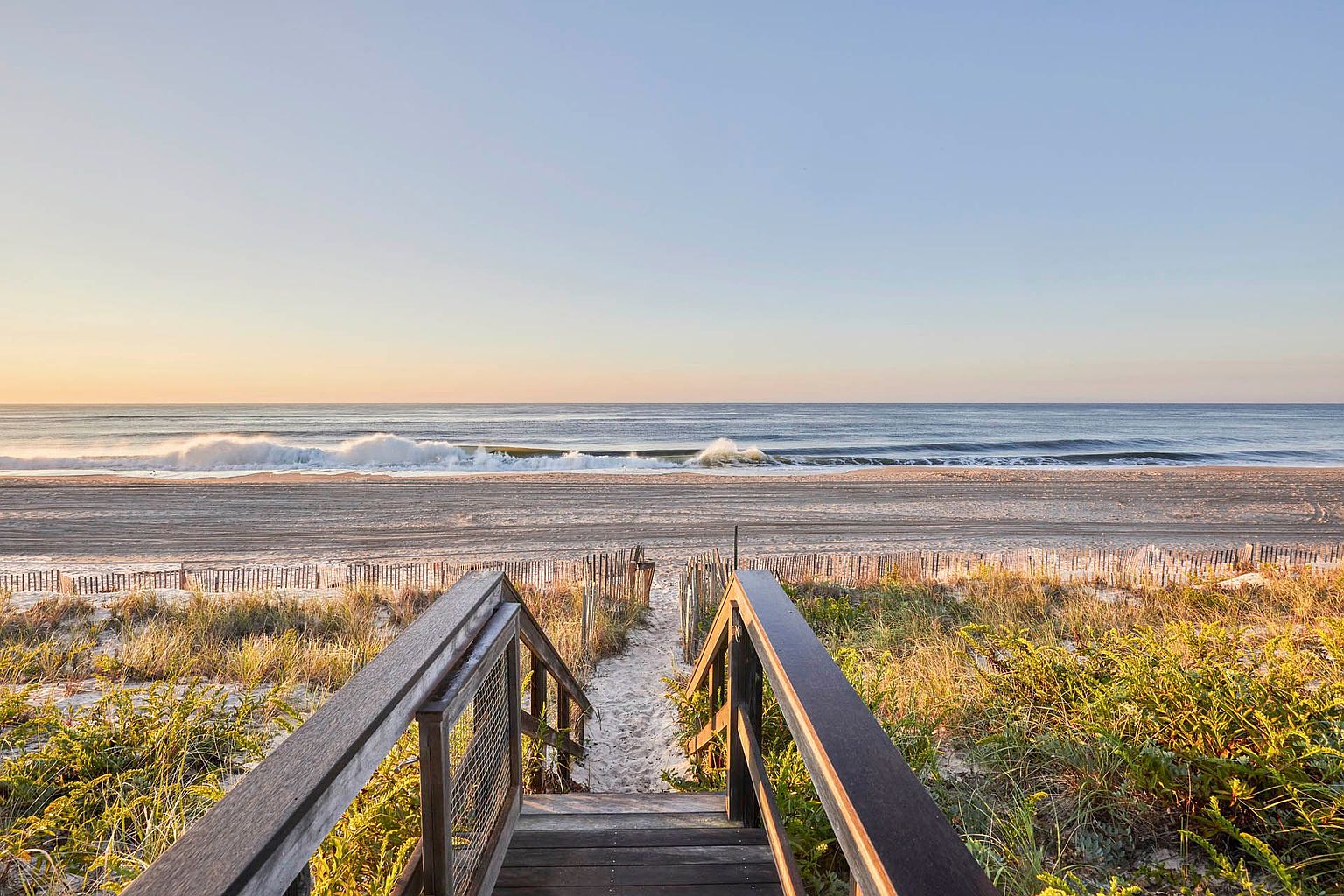 This image showcases a serene beach scene viewed from a wooden staircase leading down to the sand. The foreground features lush dune grass and a wooden fence, adding texture and depth. The ocean waves gently roll onto the shore under a clear sky, creating a peaceful and inviting atmosphere.