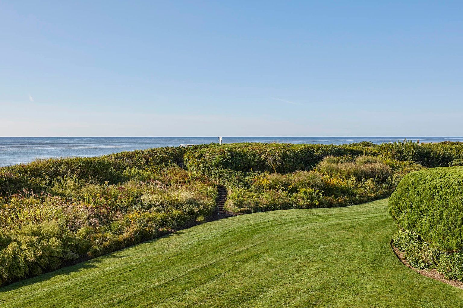 This image showcases a beautifully maintained yard and garden leading towards the ocean. A lush green lawn slopes gently towards a dense, natural landscape of shrubs and grasses, creating a seamless transition to the coastal environment. A small path leads through the vegetation towards the ocean, suggesting private access to the waterfront.