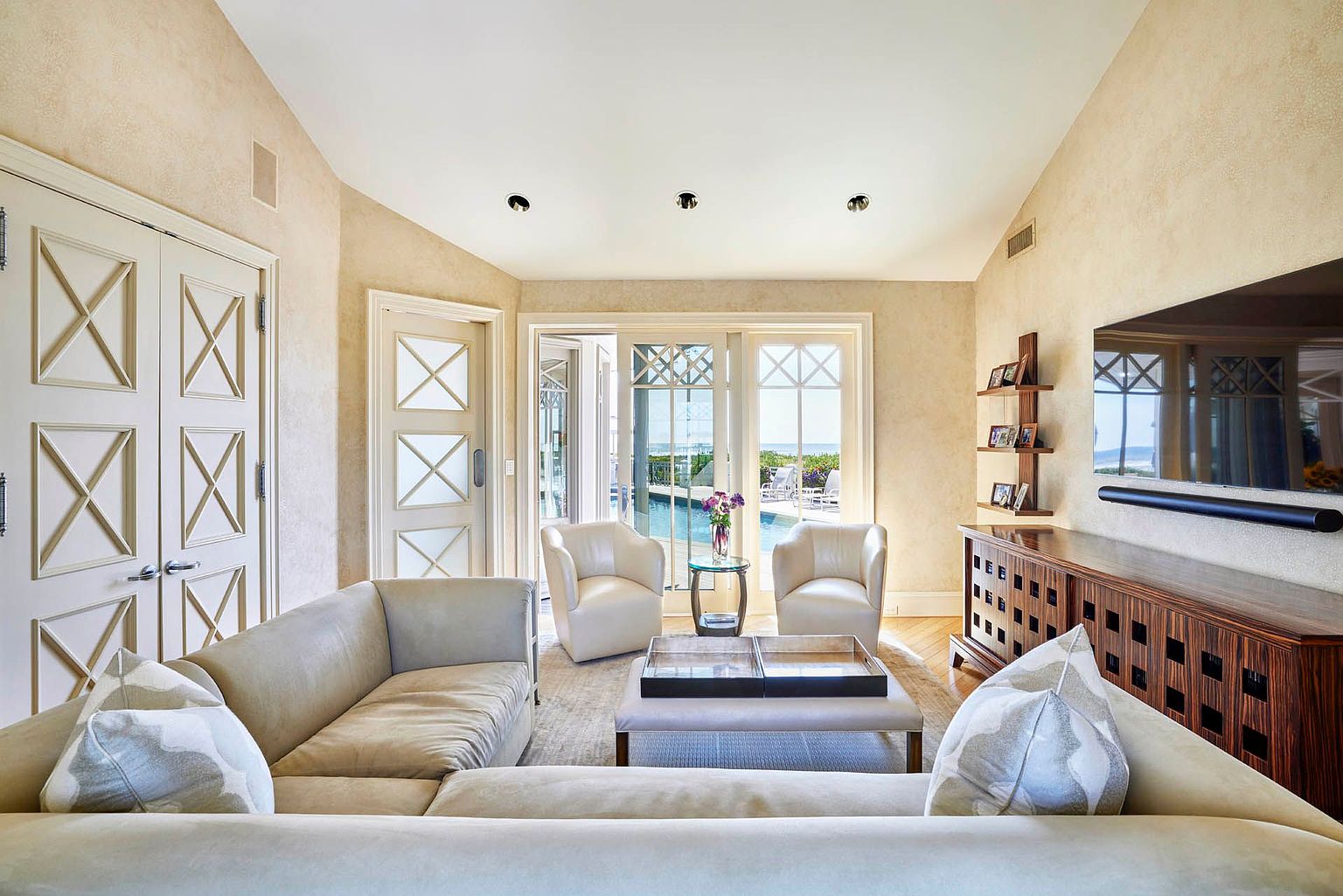This is an interior shot of a luxurious living room featuring a neutral color palette with beige walls and light-colored furniture. The room has two armchairs facing a set of glass doors that lead to an outdoor pool area. A large sectional sofa is in the foreground, and a wooden cabinet with a flat-screen TV is on the right side of the room, creating a comfortable and elegant living space.