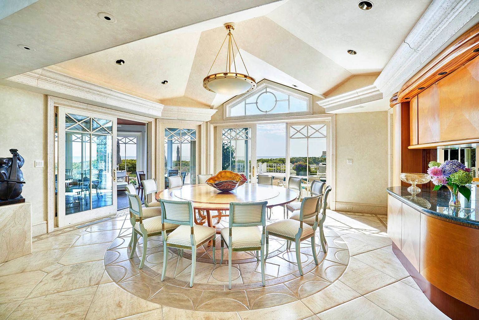 This is a bright and elegant dining room featuring a large round table surrounded by upholstered chairs. The room is well-lit with natural light streaming through large windows and a statement chandelier overhead. The flooring is a decorative tile pattern, adding to the room's sophisticated ambiance.