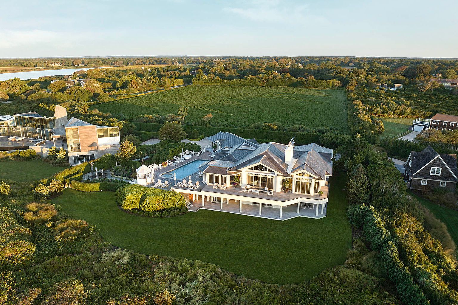 This aerial shot showcases a luxurious estate featuring a large, modern house with a gray roof and a spacious deck overlooking a well-manicured lawn. A swimming pool is visible, surrounded by lounge chairs, and the property is bordered by lush greenery and a large field. The view extends to a distant body of water and surrounding trees, creating an impression of privacy and exclusivity.