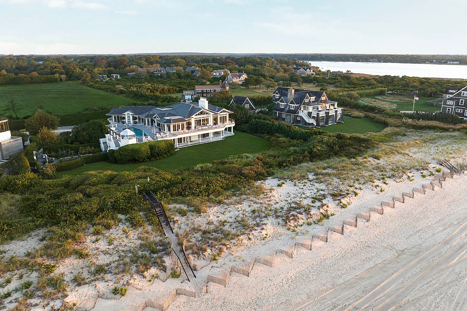 This aerial shot showcases a luxurious beachfront property featuring a grand, multi-story house with a pool and expansive deck. The manicured lawn extends to the dunes, leading to a pristine sandy beach with wooden walkways. The overall impression is one of opulence and serene coastal living.