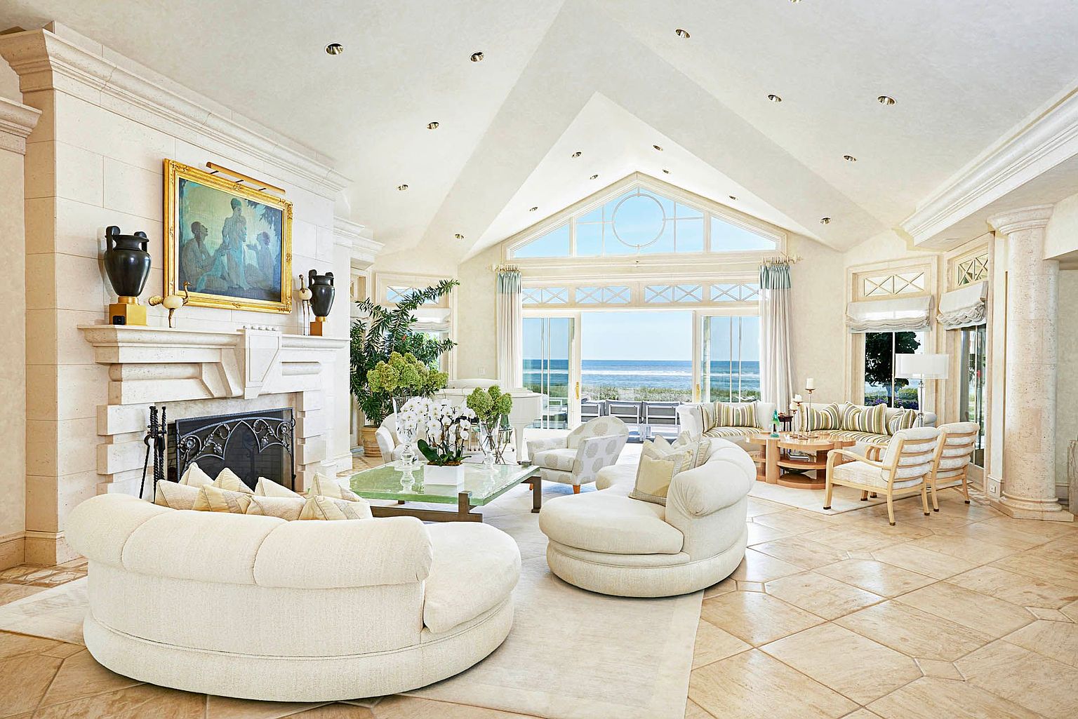 This is a luxurious living room featuring a high vaulted ceiling with recessed lighting and a large window overlooking the ocean. The room is furnished with two round white sofas, a glass coffee table, and several armchairs. A stone fireplace with decorative urns adds a touch of elegance, and the overall style is bright, airy, and sophisticated.