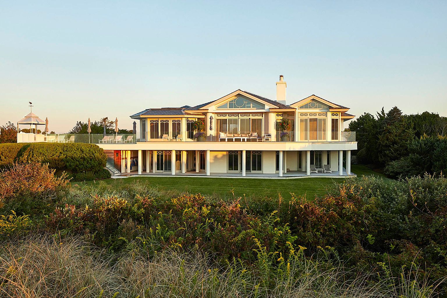 This is a rear view of a large, luxurious two-story house with a sprawling green lawn. The house features a covered patio supported by white columns, a second-story balcony with glass railings, and multiple large windows offering ample natural light. The surrounding landscape includes lush greenery and tall grasses, creating a serene and private setting.