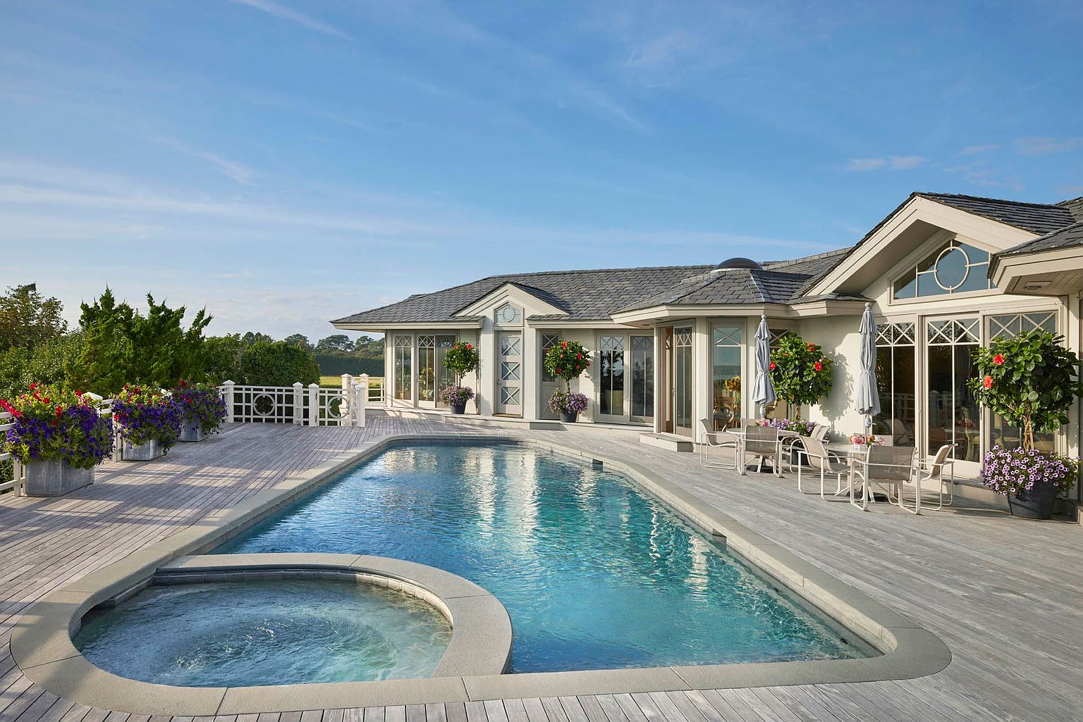 This image showcases a luxurious backyard featuring a swimming pool and spa. The pool is surrounded by a wooden deck, and the house has large windows and doors that open onto the deck. The overall impression is one of relaxation and luxury, perfect for entertaining or enjoying a quiet afternoon.