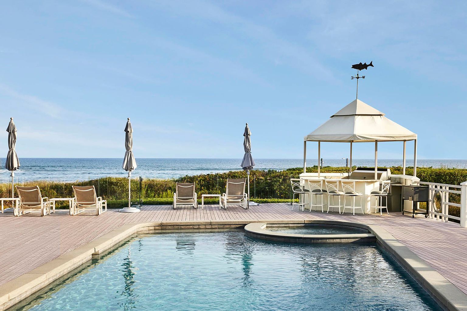 This exterior shot showcases a luxurious pool and spa area, complete with lounge chairs, umbrellas, and a covered outdoor bar. The pool is surrounded by a wooden deck, and the ocean is visible in the background, creating a serene and upscale atmosphere. The presence of a weather vane adds a unique architectural detail.