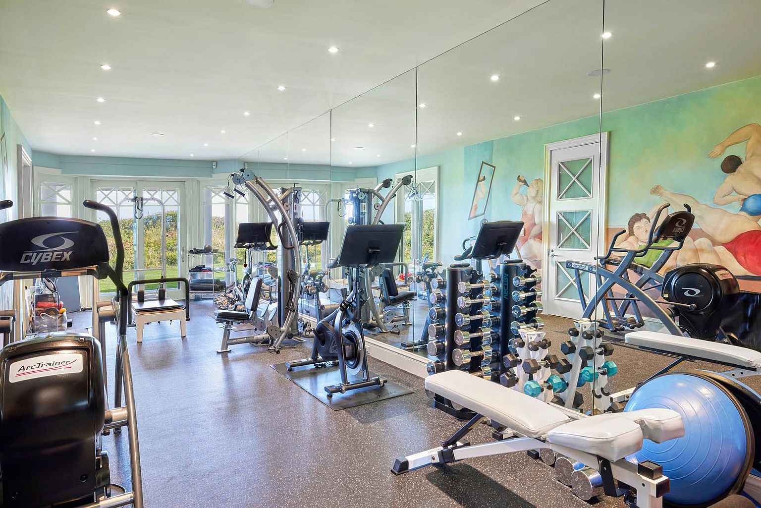 This is a spacious home gym featuring a variety of exercise equipment, including treadmills, weight machines, and free weights. A large mirror covers one wall, enhancing the sense of space and allowing for form checks. The room is well-lit with recessed lighting and natural light from the windows, creating an inviting atmosphere for workouts.