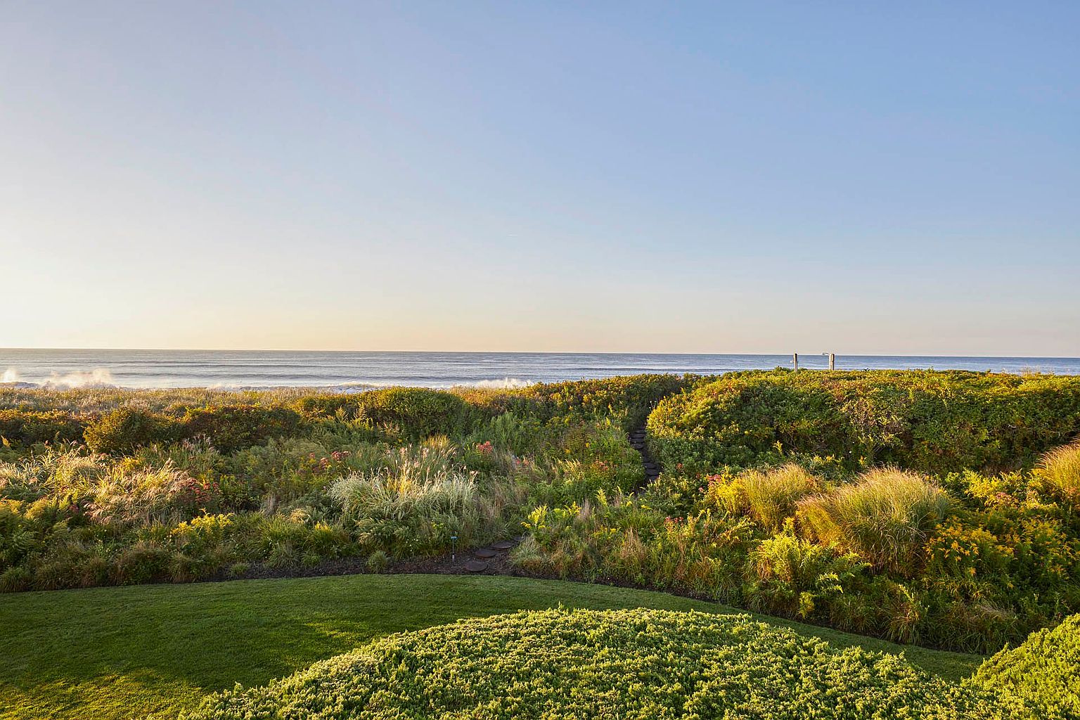 This exterior shot showcases a lush yard and garden leading to the ocean. The foreground features manicured green lawn and dense shrubbery, transitioning into a wilder garden with tall grasses and flowering plants. In the background, the ocean meets the horizon under a clear blue sky, creating a serene and inviting atmosphere.
