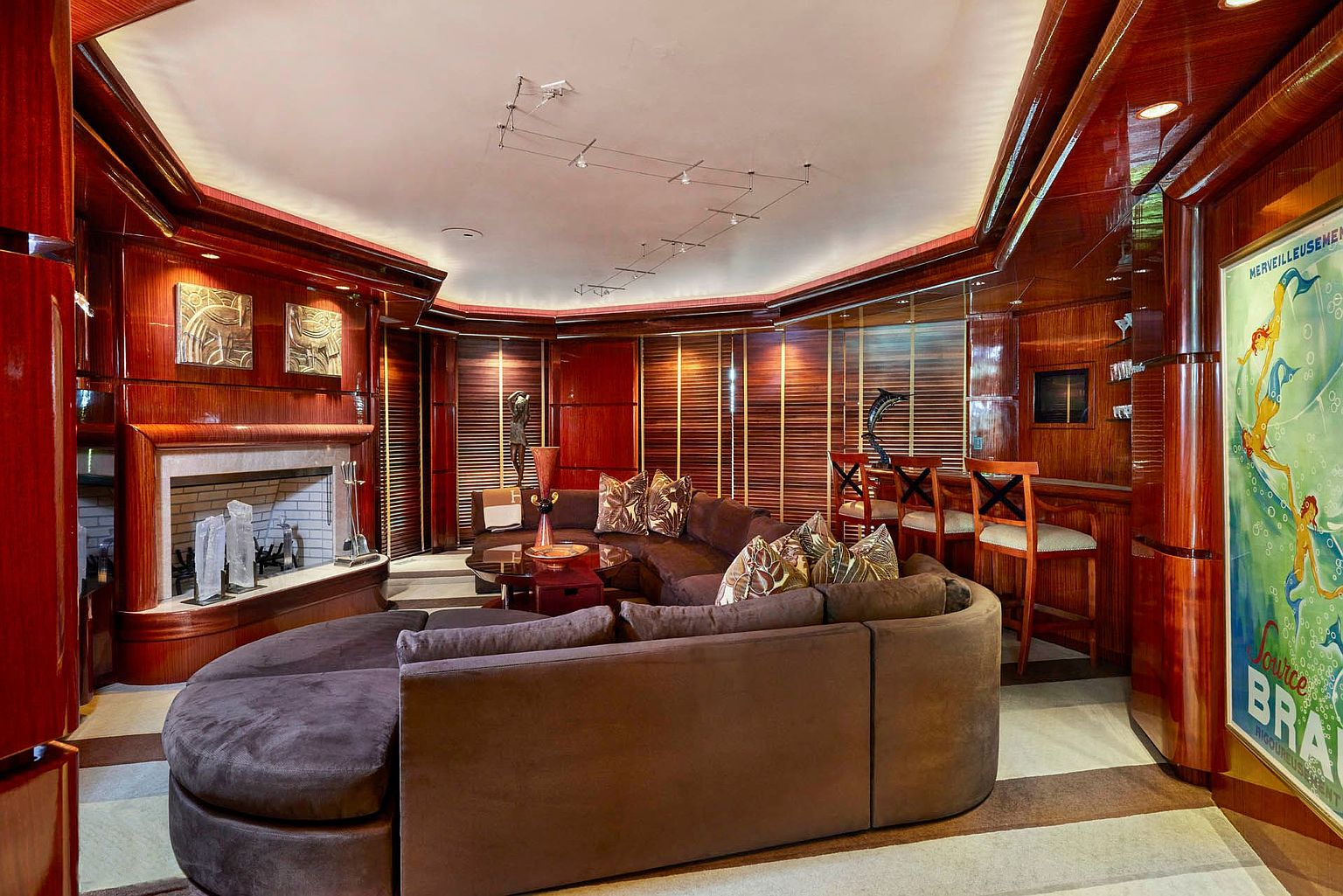 This is an interior shot of a luxurious living room, possibly on a yacht, featuring rich wood paneling and a large, curved brown sofa. A fireplace with decorative elements adds a focal point, while a bar area with stools is visible in the background. The room exudes a sophisticated and opulent atmosphere.