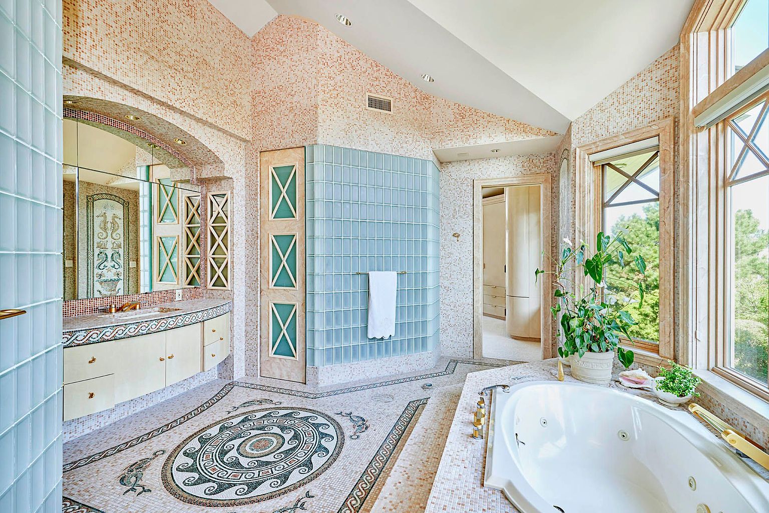 This is a luxurious primary bathroom featuring mosaic tile walls and flooring with intricate patterns. A large jacuzzi tub sits near a window offering natural light, while a glass block shower and vanity area with a large mirror complete the space. The overall impression is opulent and unique.