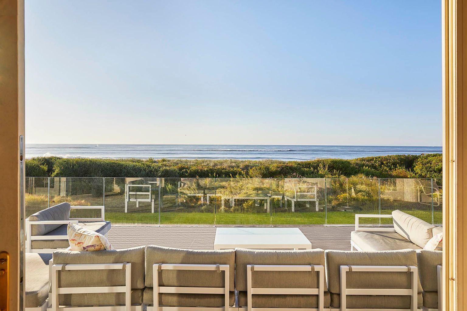 This image showcases a luxurious outdoor patio or balcony with comfortable seating arrangements, offering a serene view of the ocean. The patio features modern furniture with light-colored frames and neutral cushions, complemented by a glass railing that provides an unobstructed view of the beach and horizon. The scene evokes a sense of relaxation and upscale coastal living.