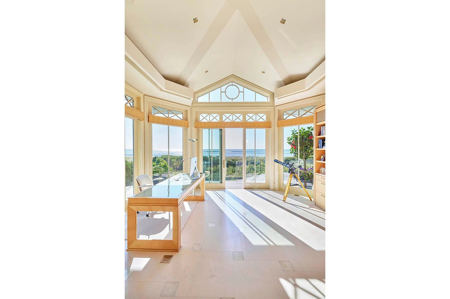 This is a bright and airy office space with a stunning ocean view. The room features a large glass-topped desk, a telescope, and a built-in bookcase. The high vaulted ceiling and large windows create a sense of spaciousness and luxury, making it an ideal workspace with a serene atmosphere.