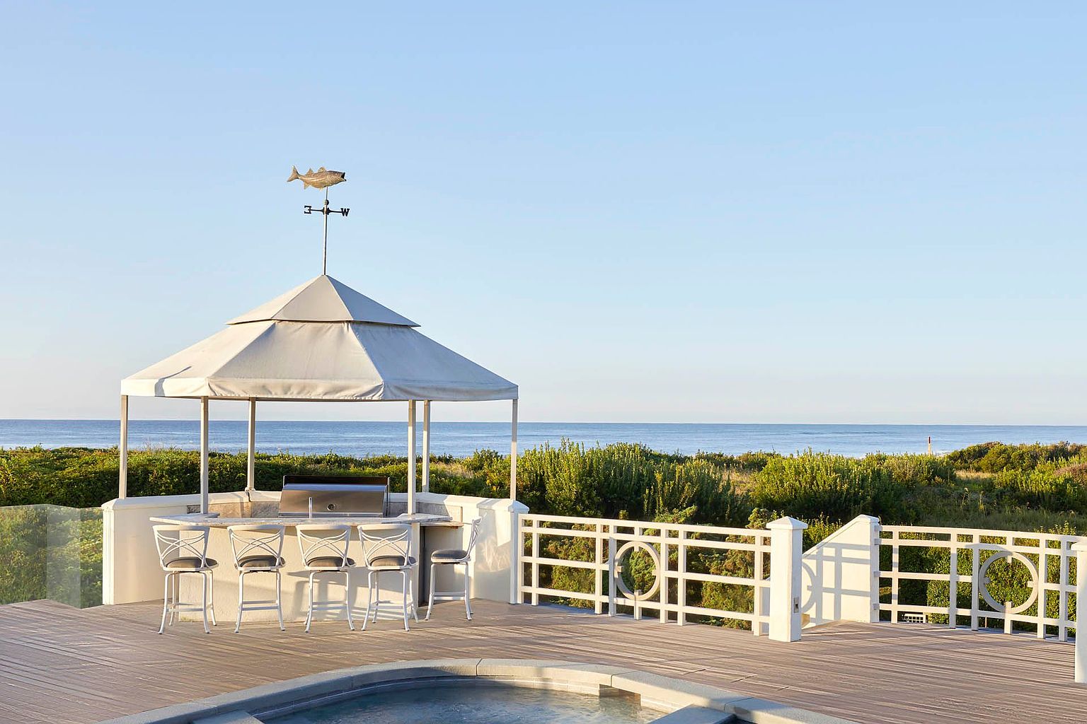 This image showcases a luxurious outdoor patio/deck area with a stunning ocean view. The space features a covered outdoor kitchen with bar seating, a decorative white fence, and a built-in spa. The deck is made of wood and the overall impression is one of high-end relaxation and entertainment.