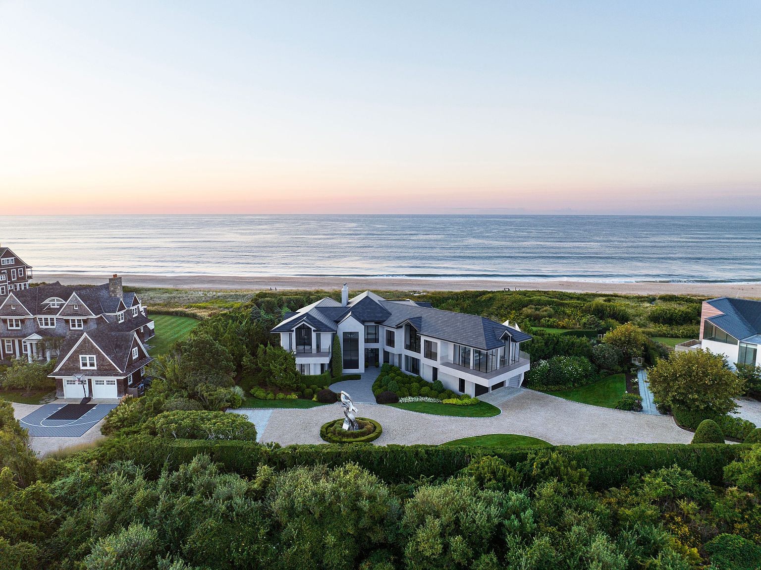 This aerial view showcases a luxurious beachfront property with meticulously landscaped grounds, including manicured lawns, mature trees, and sculpted hedges. The modern home features a light-colored exterior, dark roof, and a circular driveway with a central sculpture, all set against the backdrop of the ocean and a clear sky. The image conveys a sense of exclusivity and high-end coastal living.
