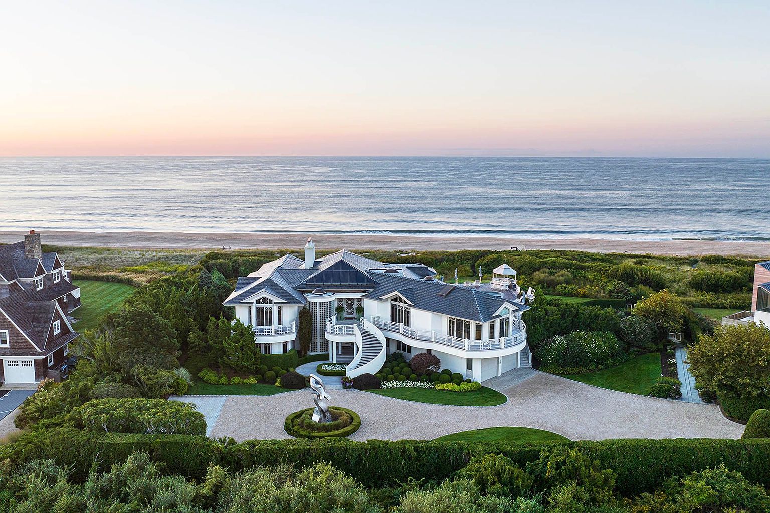 This aerial view showcases a luxurious beachfront estate with a grand circular driveway, manicured gardens, and a striking sculpture at the entrance. The house features a combination of traditional and modern architectural elements, including multiple balconies, a curved staircase, and a dark-tiled roof. The property offers direct access to a pristine sandy beach and ocean views, emphasizing its exclusive and desirable location.