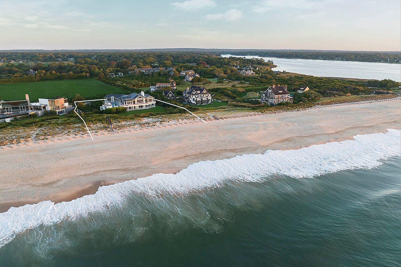 This aerial shot showcases a stunning beachfront property featuring multiple houses along a sandy coastline. The turquoise ocean meets the shore with foamy waves, while lush greenery and trees provide a scenic backdrop. The image highlights the property's prime location and luxurious coastal living.