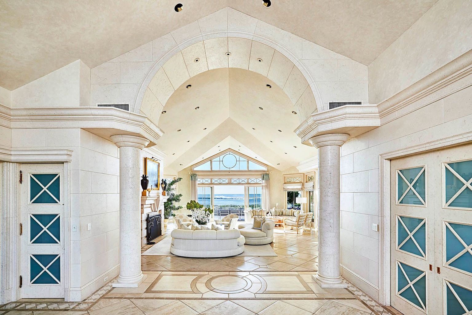 This grand living room features an open floor plan with a view of the ocean through large windows. The room is decorated in a neutral color palette with white sofas and light-colored flooring. Architectural details include arched doorways, columns, and a fireplace, creating a luxurious and inviting space.