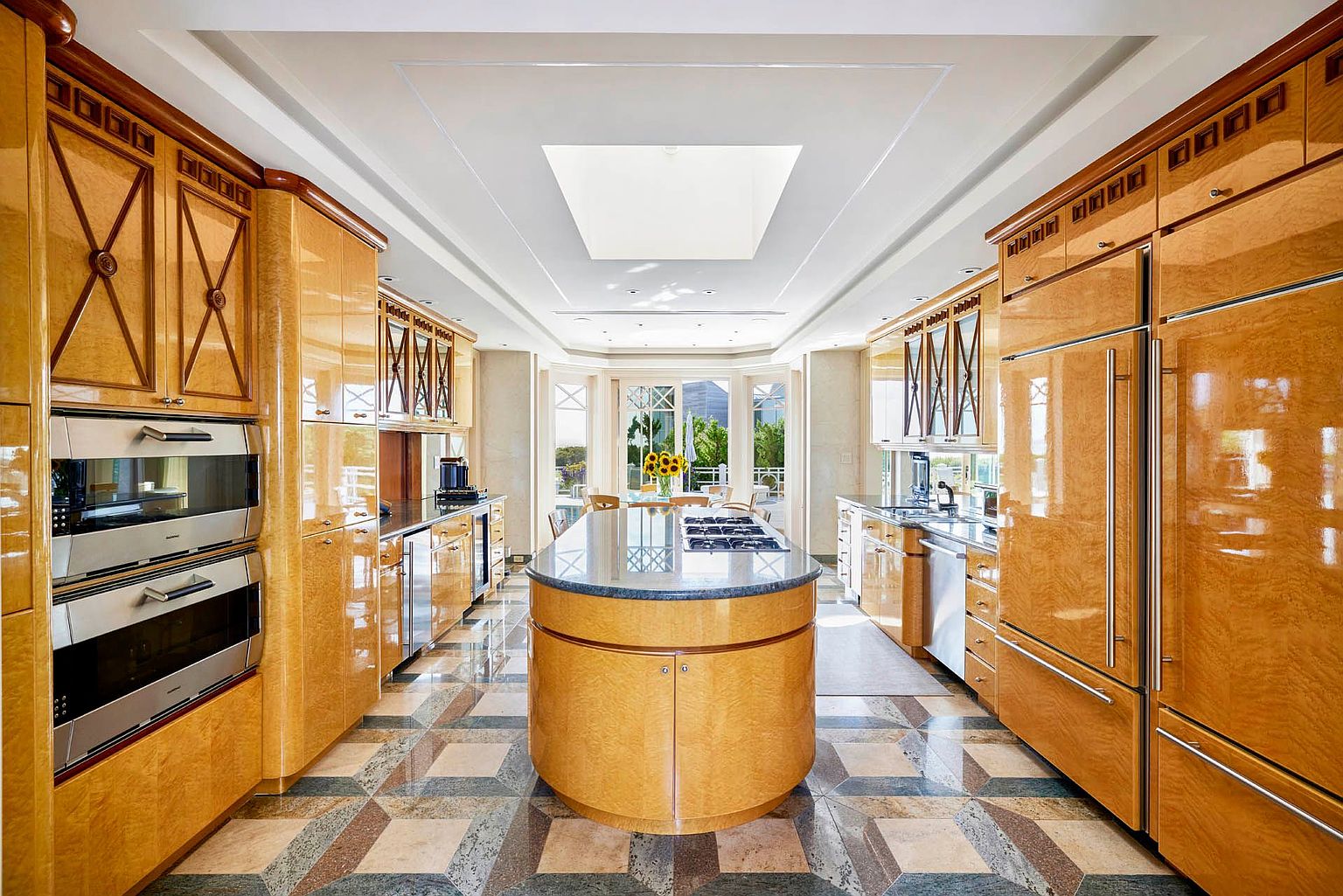 This is a high-end kitchen featuring custom wood cabinetry with decorative accents, stainless steel appliances, and a large island with a dark countertop. The floor is tiled in a geometric pattern, and a skylight provides natural light. The kitchen opens to an outdoor area through large windows, creating a bright and luxurious space.