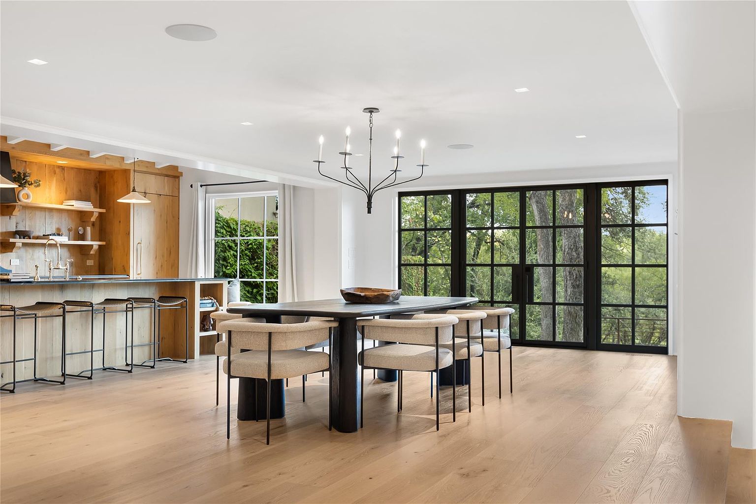 This is an interior shot of a dining room featuring a dark wood dining table with modern chairs. A chandelier hangs above the table, and large windows with black frames offer a view of the greenery outside. The room has light wood floors and white walls, creating a bright and airy atmosphere.