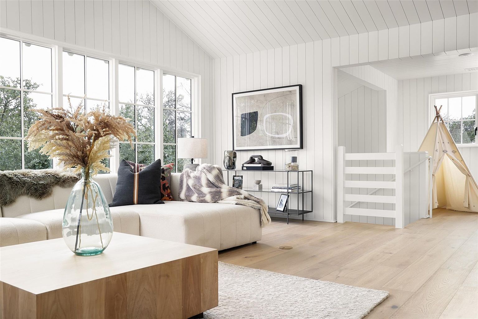 This is a bright and airy living room featuring white paneled walls and ceiling, large windows, and light wood flooring. A sectional sofa is adorned with decorative pillows and a throw blanket, while a modern coffee table sits in the foreground. The room also includes a stylish shelving unit and a playful teepee in the background, creating a welcoming and contemporary atmosphere.