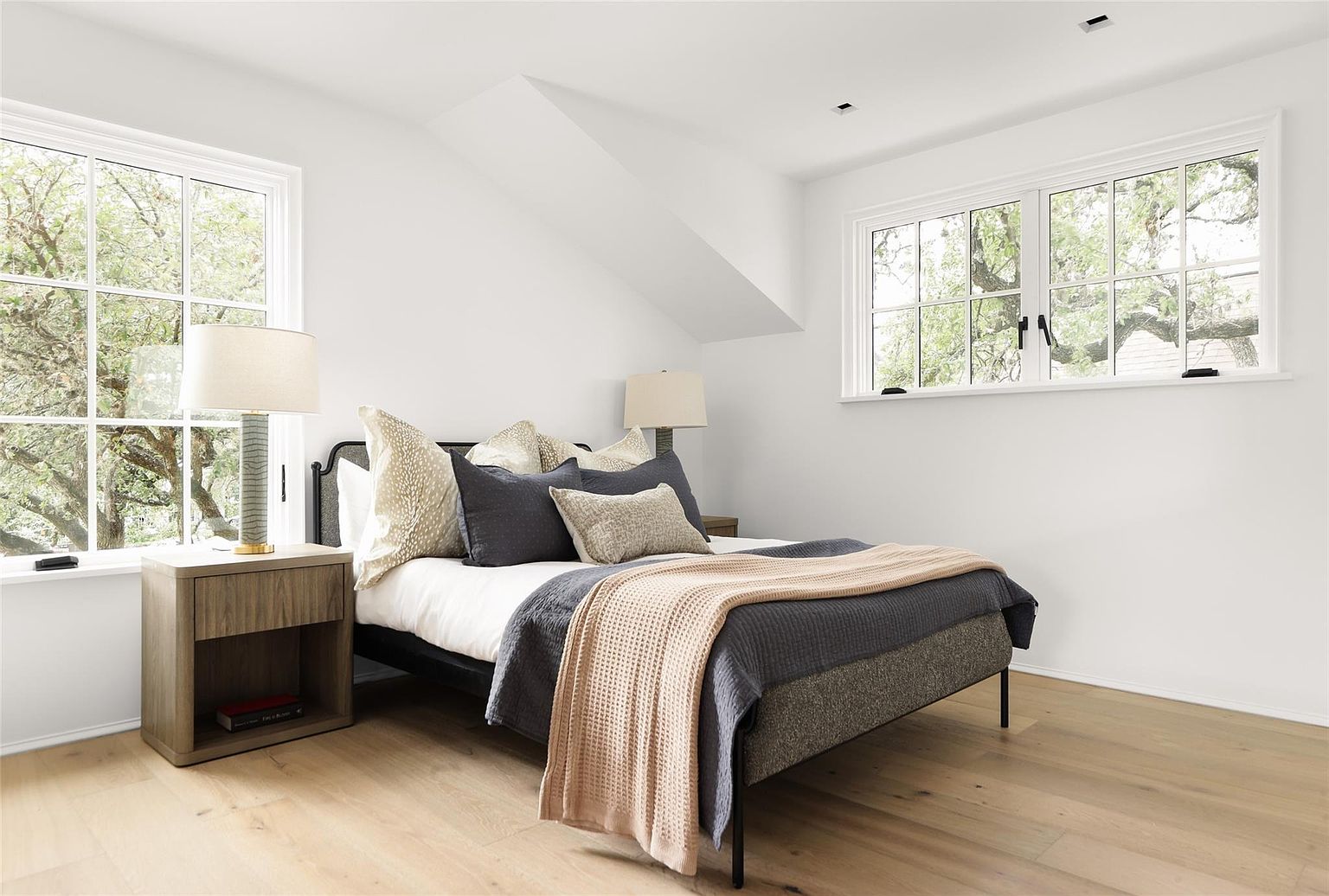 This is a bright and airy bedroom featuring a bed with neutral-toned bedding and pillows, flanked by a wooden nightstand with a lamp. Natural light floods the room through two large windows, highlighting the light hardwood flooring and white walls. The overall impression is clean, comfortable, and inviting.
