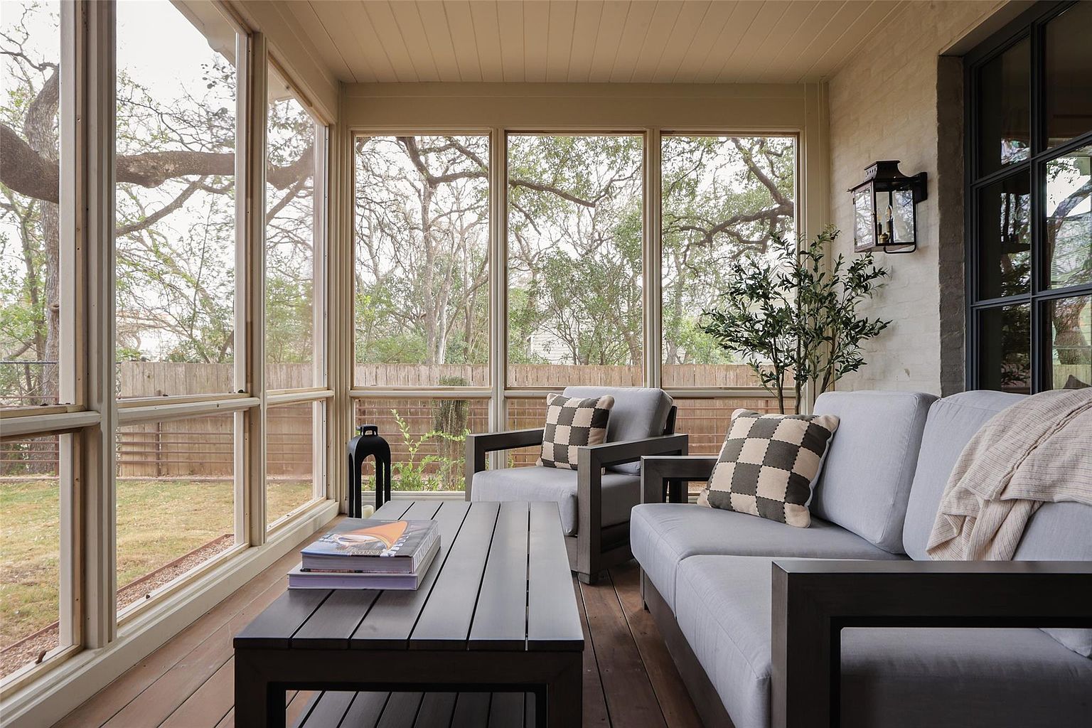 This image showcases a screened-in porch or patio area, furnished with a gray sofa, armchair, and a dark wood coffee table. Large windows offer views of the outdoor greenery, creating a seamless transition between indoor and outdoor living. The space is well-lit and appears to be designed for relaxation and entertainment.