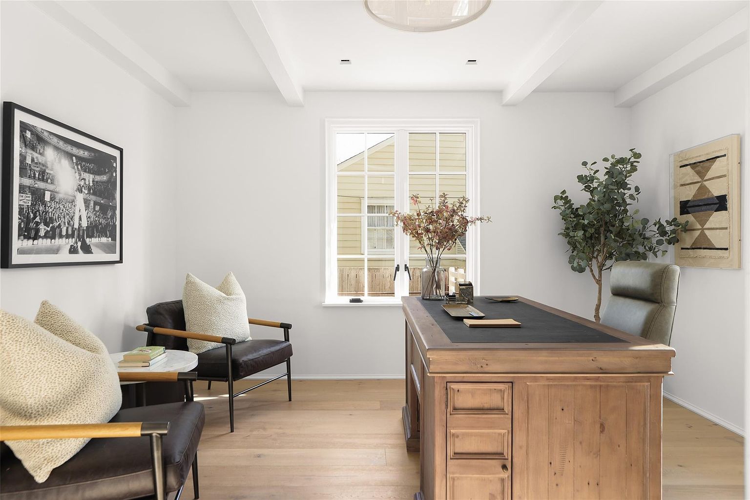 This is an interior shot of a well-lit home office. The room features a wooden desk with a black leather top, two leather chairs with light-colored cushions, and a black and white framed photograph on the wall. A large window provides natural light, and a plant adds a touch of greenery to the space.