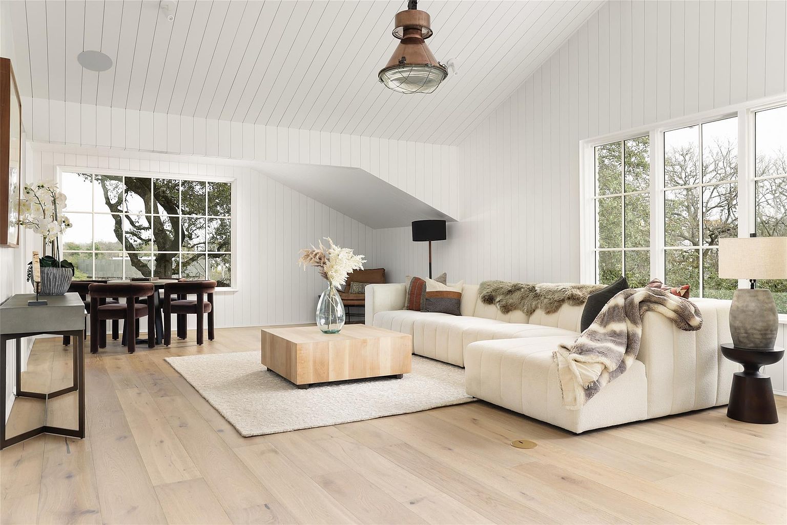 This is a bright and airy living room featuring white walls and light wood flooring. A large sectional sofa is the focal point, complemented by a light-colored area rug and a wooden coffee table. Natural light floods the space through large windows, creating a warm and inviting atmosphere.