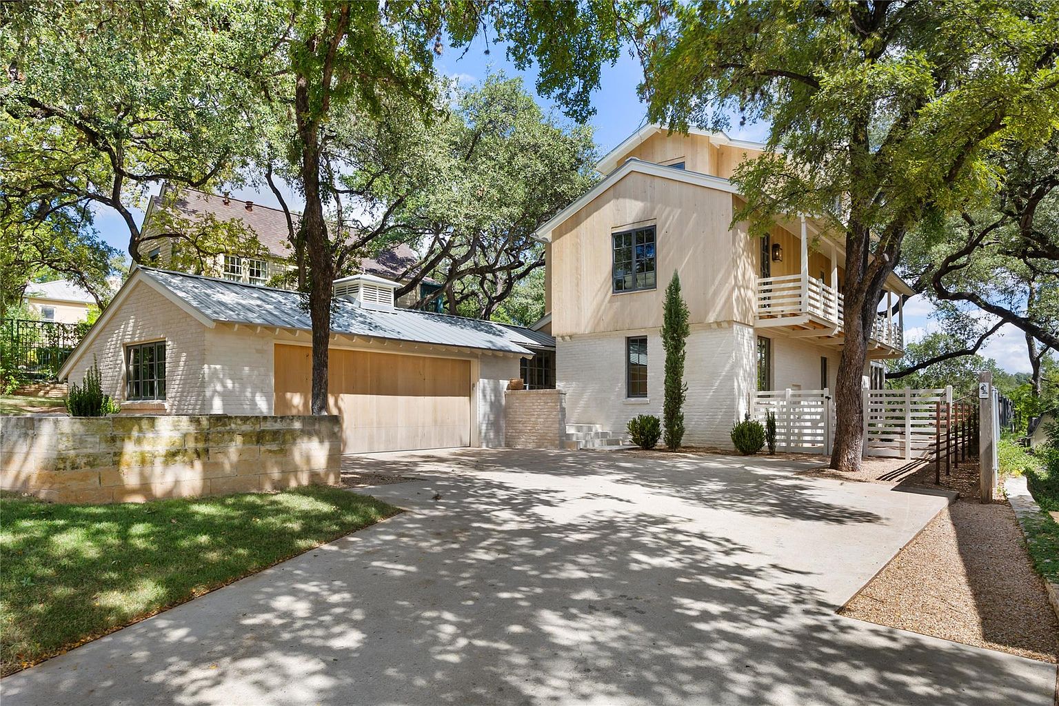 This is a front exterior view of a modern home with a light-colored brick and wood facade. The property includes a detached garage with a wooden door and a metal roof. Mature trees surround the house, providing shade and enhancing the curb appeal. A concrete driveway leads to the garage and the house, offering ample parking space.