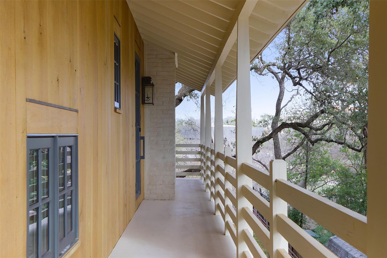 This image showcases a charming outdoor balcony or patio area. The space features a white railing and support beams, offering a bright and airy feel. The flooring is a light neutral color, and the surrounding trees provide a sense of privacy and connection to nature, enhancing the appeal of this outdoor living space.