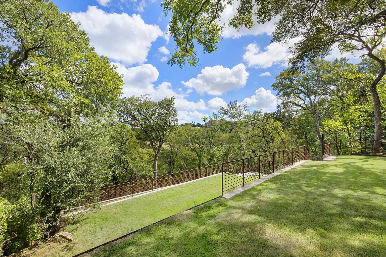 This image showcases a beautifully landscaped yard with lush green grass and mature trees. A modern metal fence runs along the perimeter, providing a clean and stylish boundary. The view extends into a wooded area, creating a sense of privacy and tranquility, with a blue sky and puffy white clouds above.