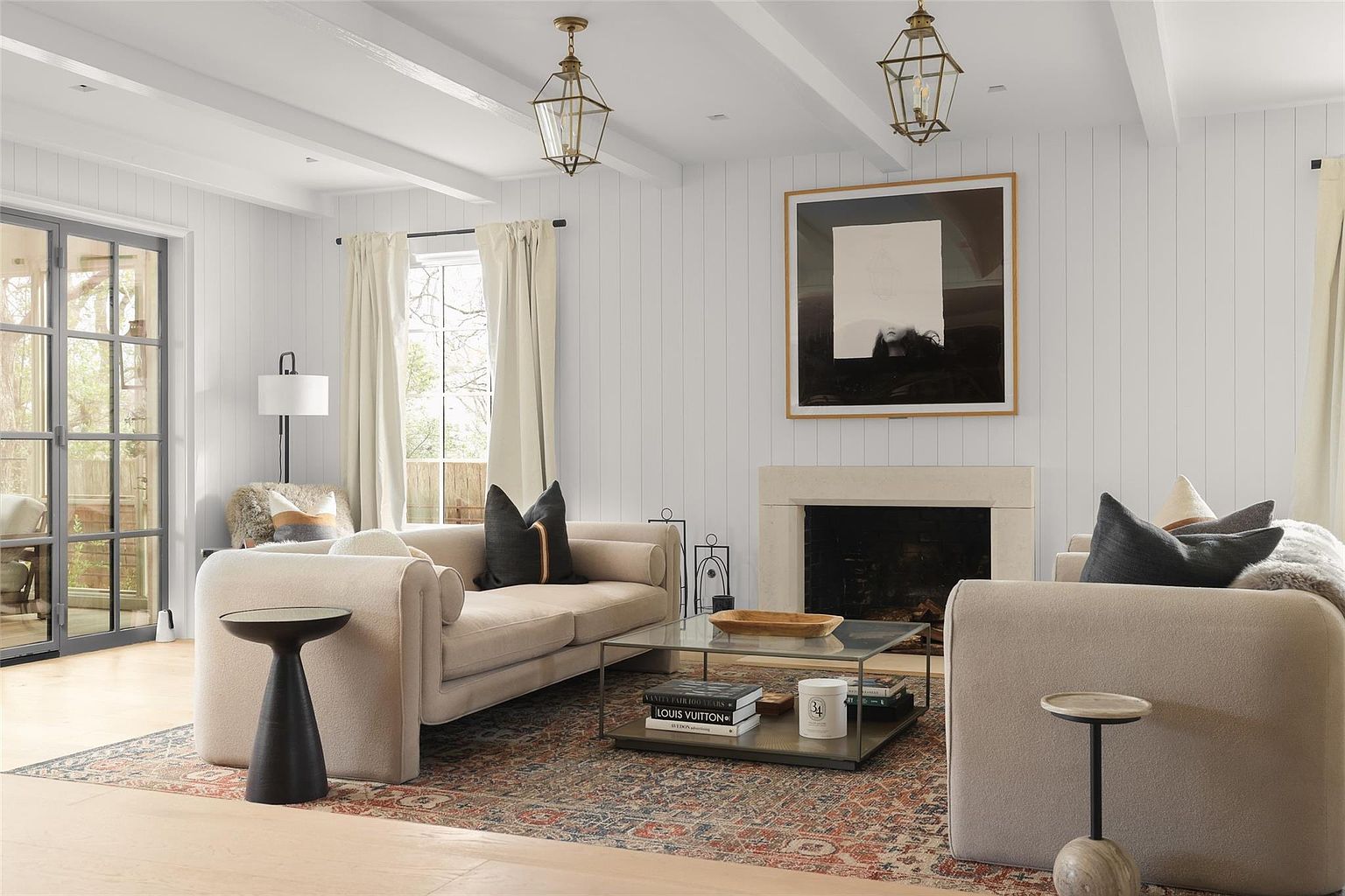 This is an interior shot of a living room featuring a neutral color palette with white walls and light-colored furniture. The room is well-lit with natural light coming through the windows and complemented by pendant lighting. A fireplace with a stone surround adds a focal point, and an area rug anchors the seating arrangement, creating a cozy and inviting atmosphere.