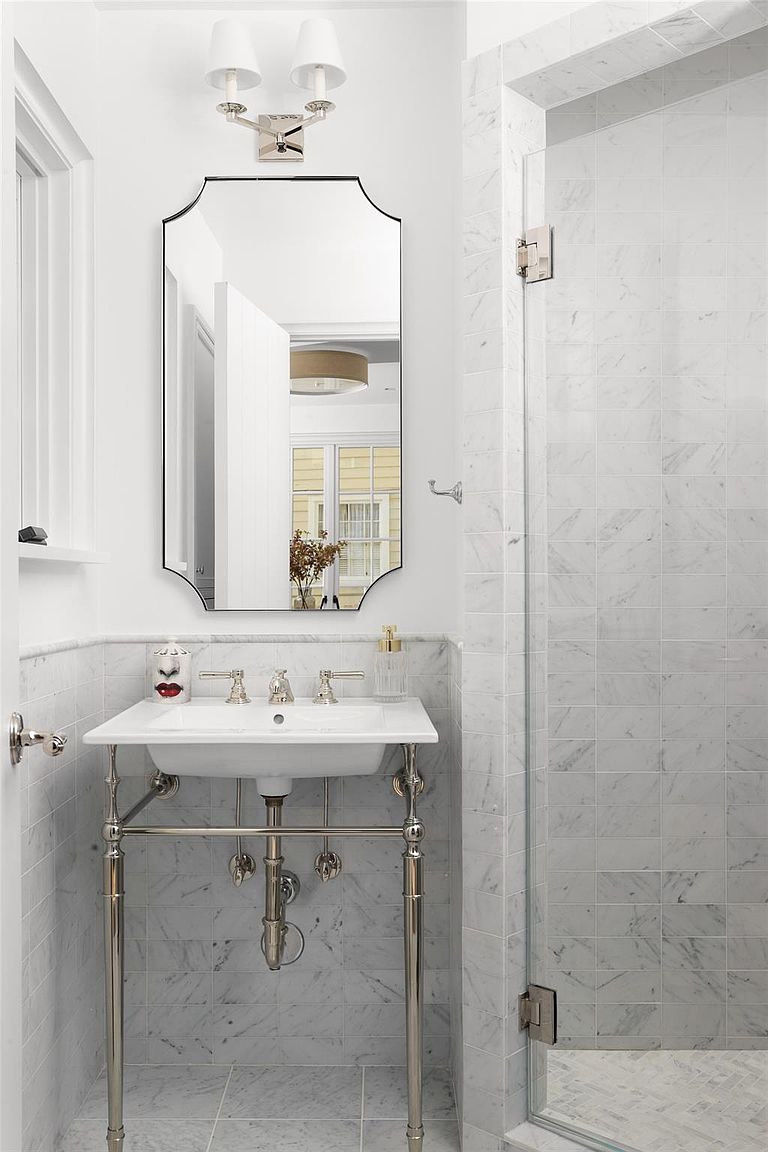 This is a well-lit bathroom featuring a pedestal sink with chrome legs and fixtures, set against a backdrop of marble tile. A frameless shower enclosure with marble tile walls is visible to the right. The overall impression is clean, classic, and luxurious.