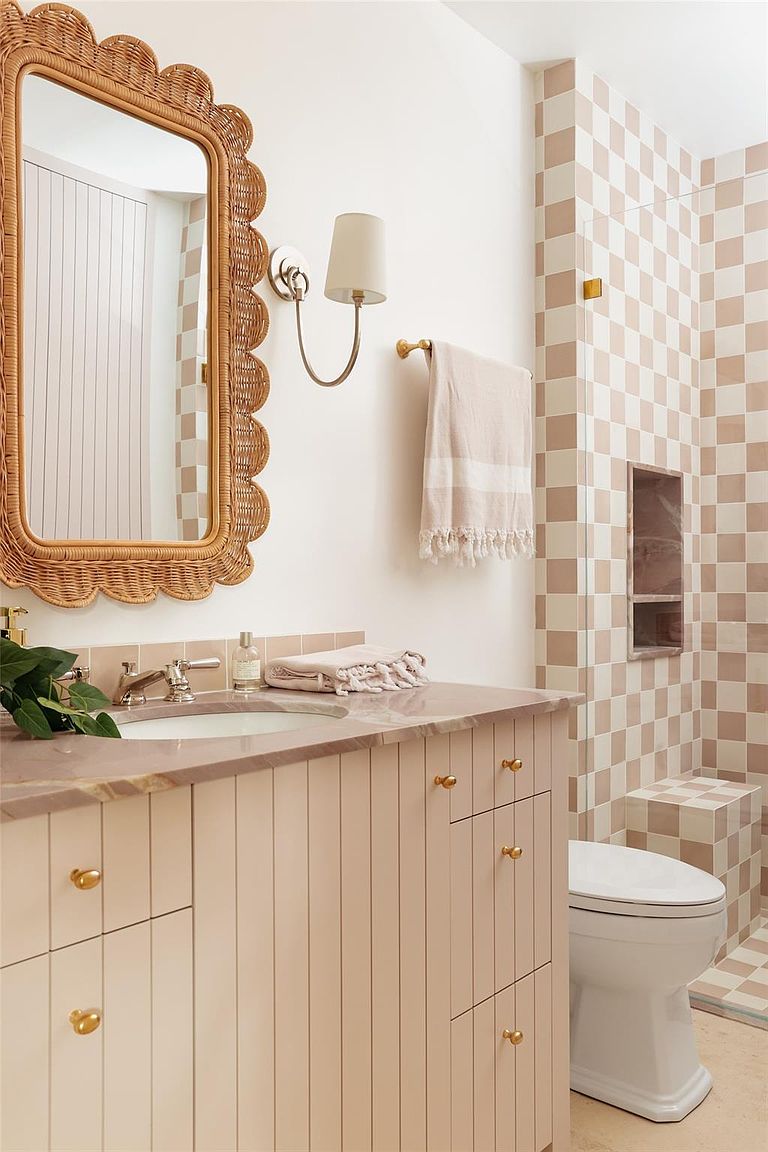 This bathroom features a charming design with a rattan-framed mirror and a vanity with vertical paneling and gold hardware. The shower area is tiled in a checkered pattern, adding a playful touch. A soft, neutral color palette creates a warm and inviting atmosphere.