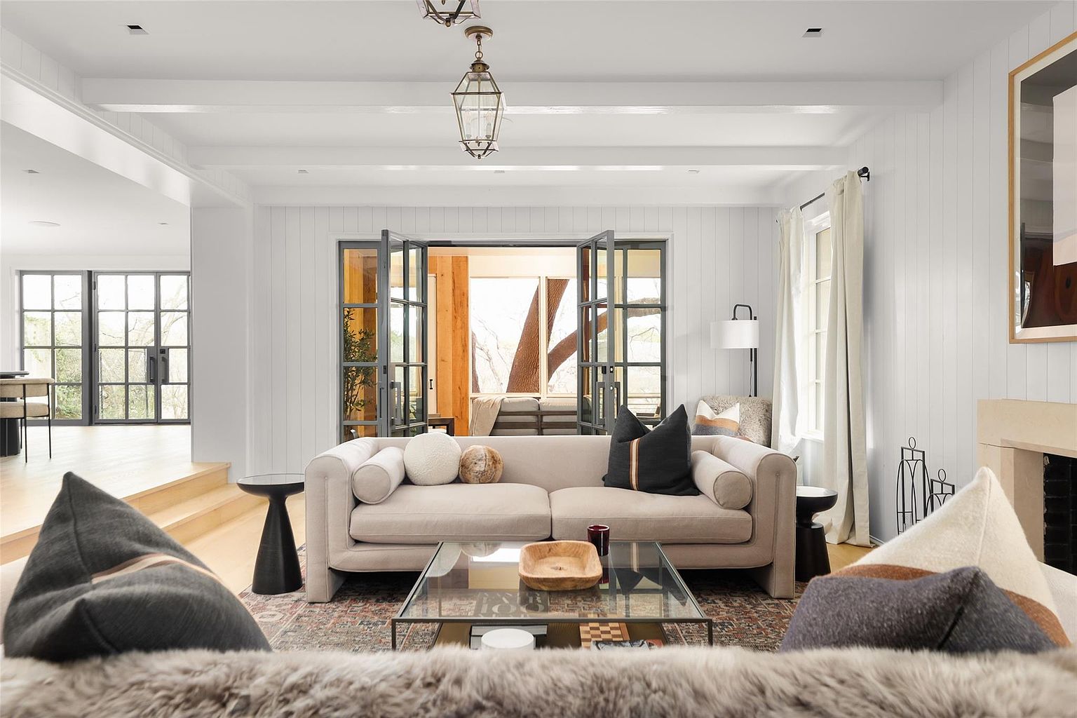 This is a bright and airy living room featuring white walls, exposed beams, and large windows/doors that provide ample natural light. A comfortable beige sofa is centered in the room, complemented by a glass coffee table and stylish side tables. The room exudes a modern, yet cozy atmosphere, perfect for relaxation and entertaining.