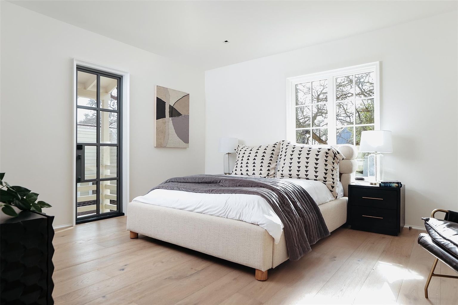 This is a bright and airy primary bedroom featuring a neutral color palette. The room is well-lit with natural light from a window and a glass door, complemented by a modern bed with a textured headboard and decorative pillows. A dark wood nightstand and artwork add contrast to the light walls and wood flooring, creating a serene and stylish atmosphere.