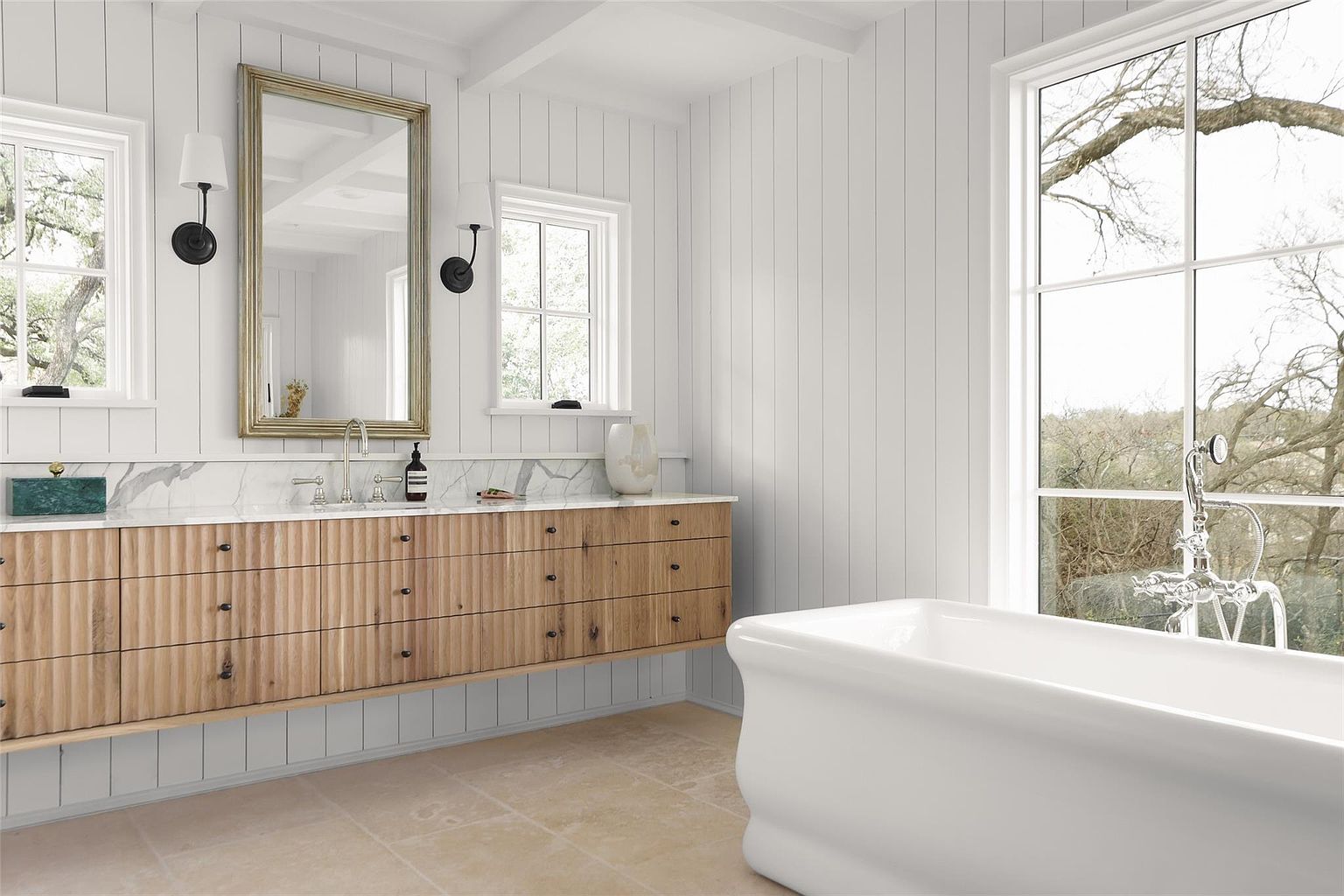 This is a bright and airy primary bathroom featuring a modern aesthetic with white paneled walls and a light wood vanity with a marble countertop. A large mirror hangs above the vanity, flanked by sconces, and a freestanding bathtub sits near a large window offering natural light and a view of the outdoors. The overall impression is clean, luxurious, and spa-like.