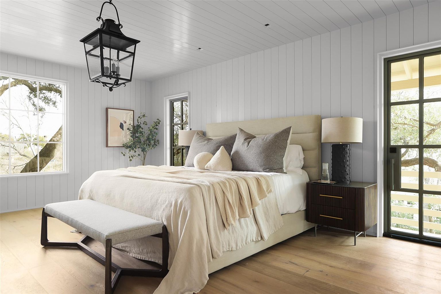 This is a bright and airy primary bedroom featuring a neutral color palette. The room is well-lit with natural light from multiple windows, highlighting the hardwood floors and paneled walls. A large bed with a padded headboard is the focal point, complemented by a dark wood nightstand and a stylish bench at the foot of the bed.