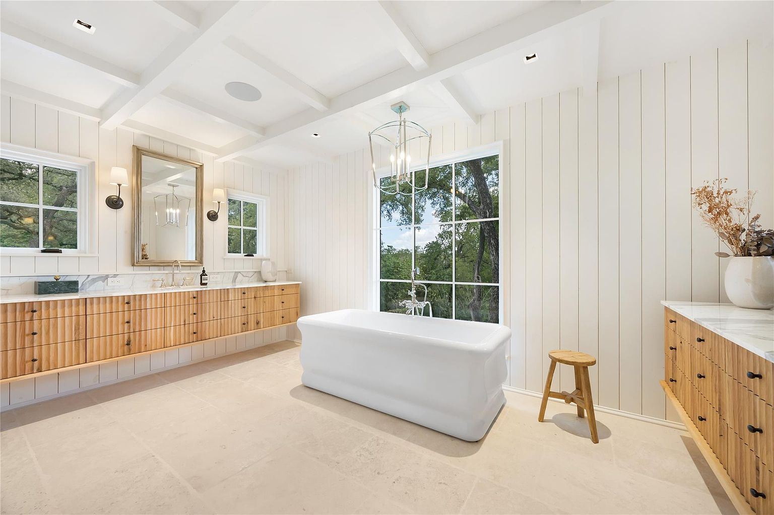 This is a bright and airy primary bathroom featuring a freestanding white bathtub positioned in front of a large window offering a view of lush greenery. The room is adorned with light wood vanities topped with marble countertops, complemented by white paneled walls and a coffered ceiling. A stylish chandelier hangs above the tub, adding a touch of elegance.