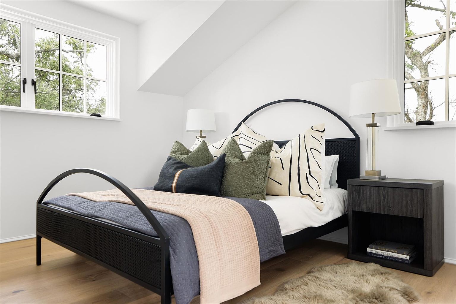 This is a cozy bedroom featuring a black metal bed frame with a woven headboard. The bed is adorned with a variety of pillows in shades of green, navy, and cream, along with a gray quilt and a light pink throw. A dark wood nightstand with a lamp sits beside the bed, and a faux fur rug adds warmth to the hardwood floor. Natural light streams in through two windows, creating a bright and inviting atmosphere.