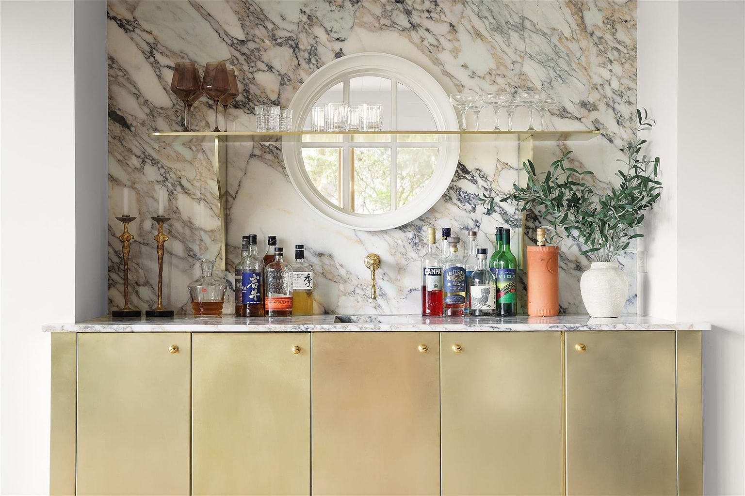 This image showcases a stylish home bar area with a marble backsplash and countertop, complemented by gold-toned cabinetry. A round mirror adds a unique architectural detail, while various bottles and glassware suggest a sophisticated entertaining space. The overall impression is one of luxury and refined taste.