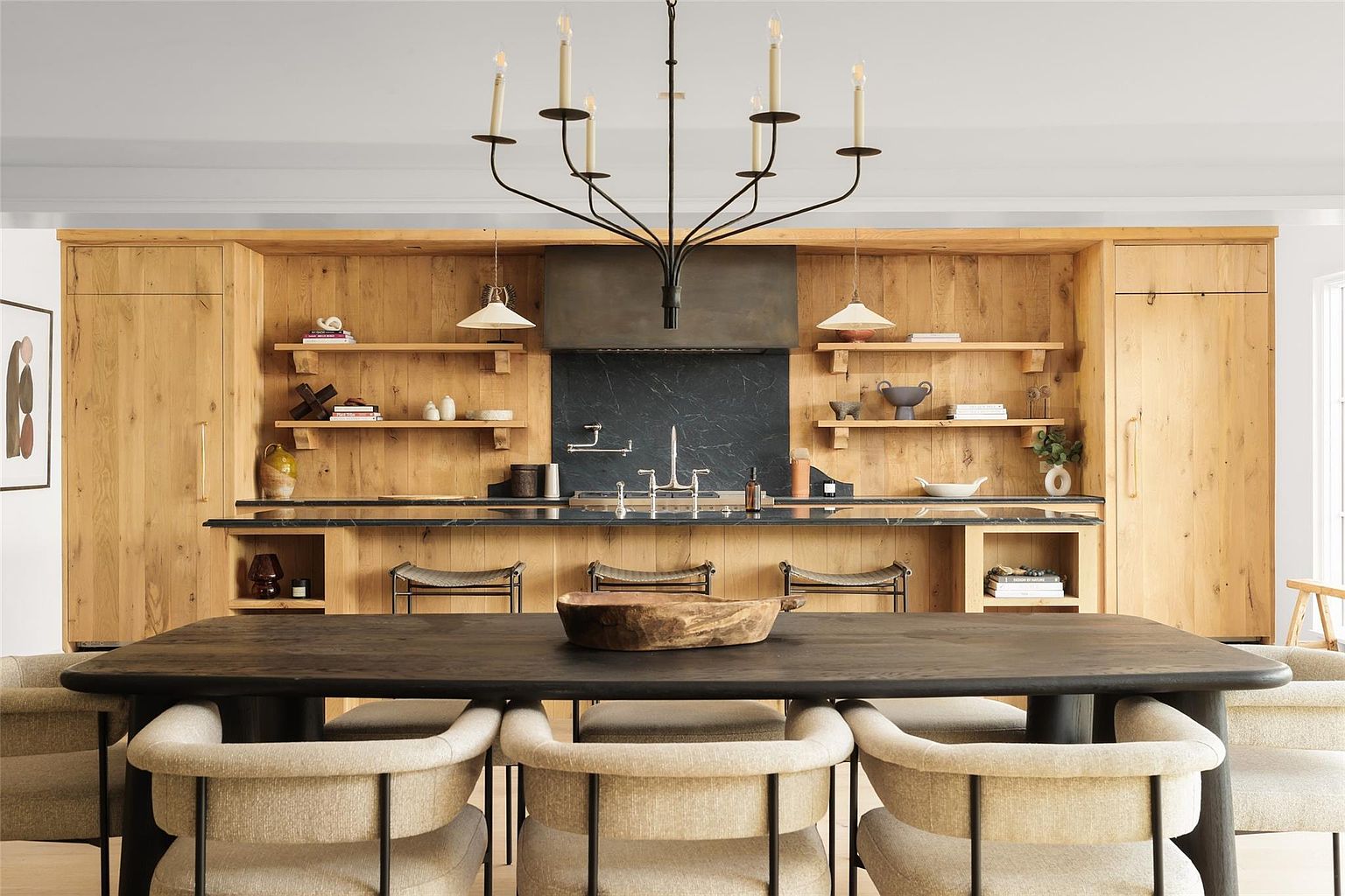 This is a well-lit kitchen featuring natural wood cabinetry and a black marble backsplash. A large island with a black countertop provides ample workspace, complemented by open shelving and stylish bar stools. A modern chandelier hangs above a dark wood dining table surrounded by upholstered chairs, creating a warm and inviting atmosphere.